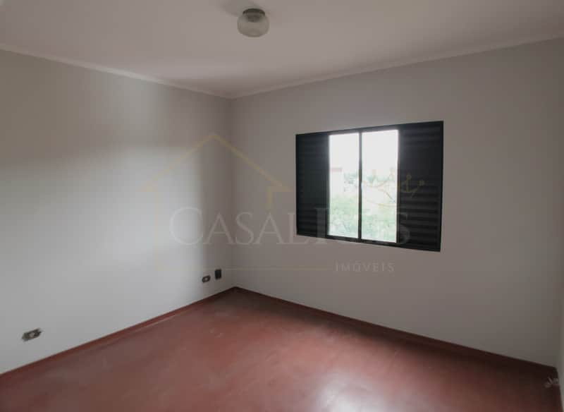 Quarto vazio com piso avermelhado e janela com venezianas escuras. Teto branco com luminária central.