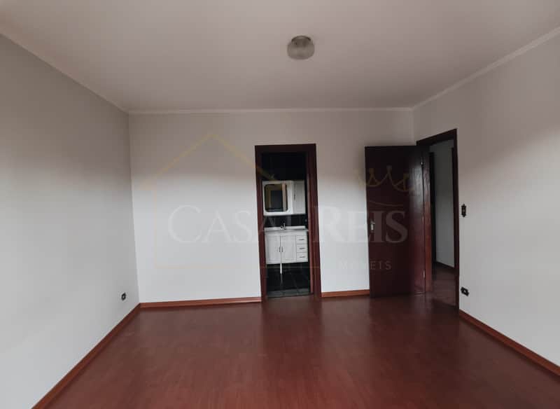 Sala ampla com piso de madeira, paredes claras e duas portas escuras. Iluminação central.