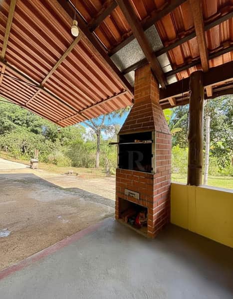 Área externa com churrasqueira de tijolos, telhado rústico e piso de cimento. Espaço amplo e arborizado.