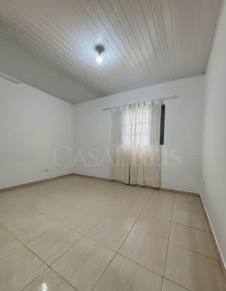 Quarto amplo e iluminado. Piso cerâmico claro, paredes brancas e teto em PVC. Janela com cortina translúcida.