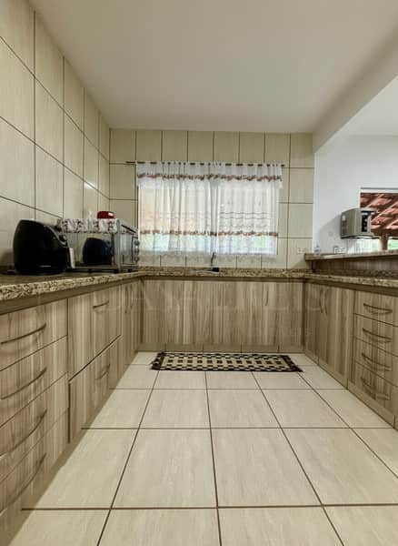 Cozinha clara com armários amadeirados, pia sob janela com cortina e piso cerâmico bege.