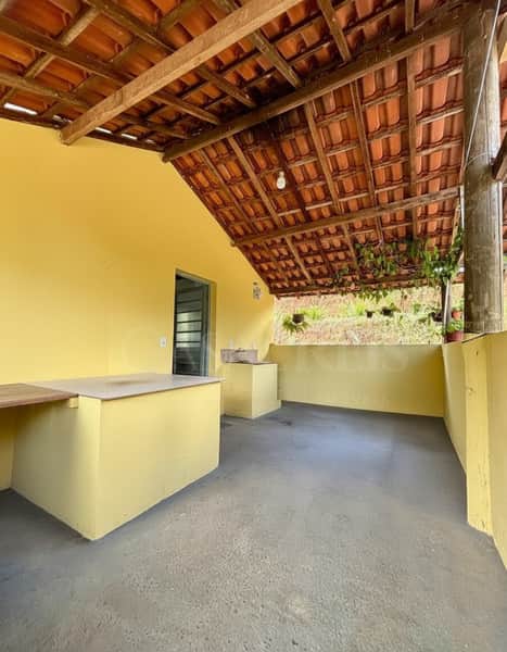 Área externa amarela com piso cinza e telhado rústico. Bancada e vegetação ao fundo.