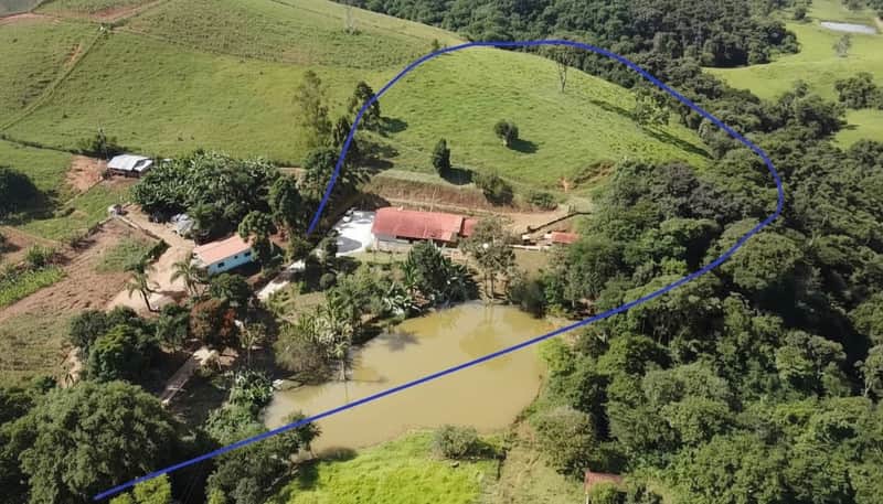 Área externa rural com lago, casa com telhado vermelho e colina verde. Vista panorâmica e arborizada.