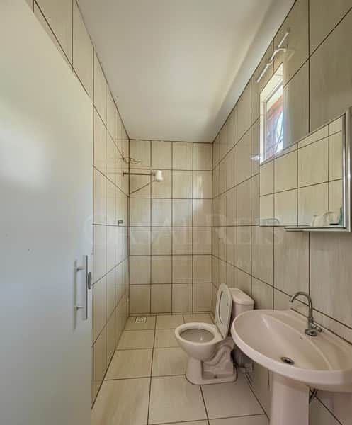 Banheiro com azulejos claros, vaso sanitário branco e pia com coluna. Espelho e janela completam o ambiente.