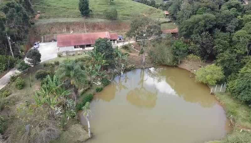 Área externa com lago, casa com telhado vermelho e vegetação exuberante. Vista panorâmica da propriedade rural.