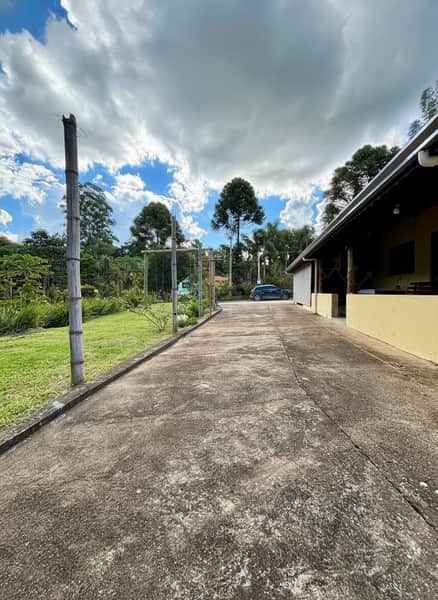 Área externa com casa amarela, gramado e árvores. Caminho de concreto leva a um carro ao fundo. Céu nublado.