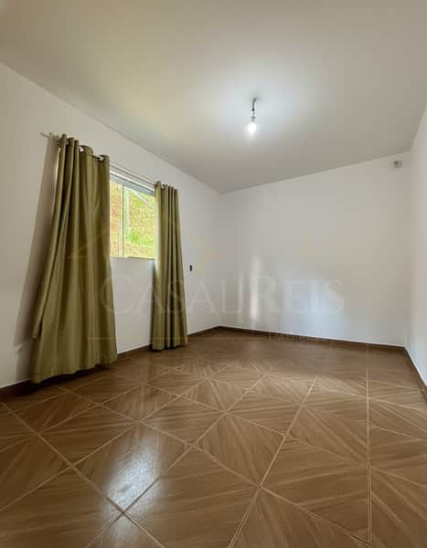 Quarto amplo com piso cerâmico, cortinas beges e janela com vista para área verde. Iluminação central.