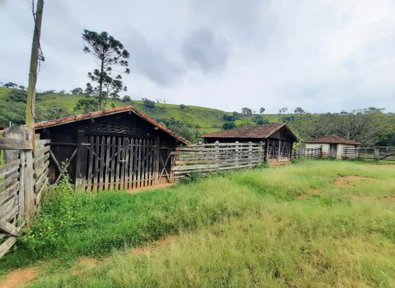 Área externa rural com currais de madeira e vegetação campestre sob céu nublado.