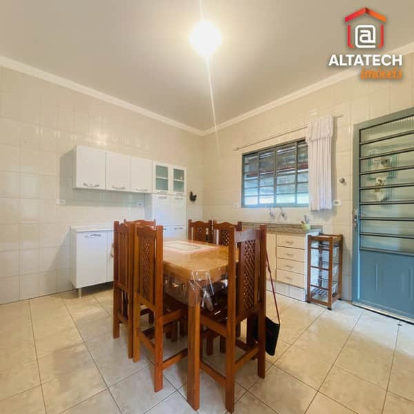 Cozinha ampla com mesa de madeira, armários brancos e piso cerâmico claro. Ambiente iluminado e arejado.