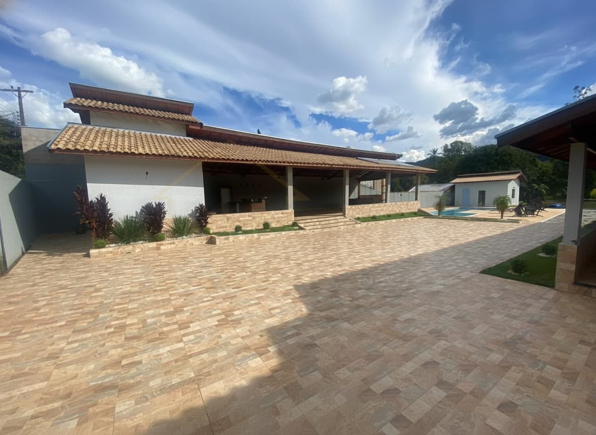 Ampla área externa com piso cerâmico, casa com varanda e piscina ao fundo. Céu azul com nuvens.