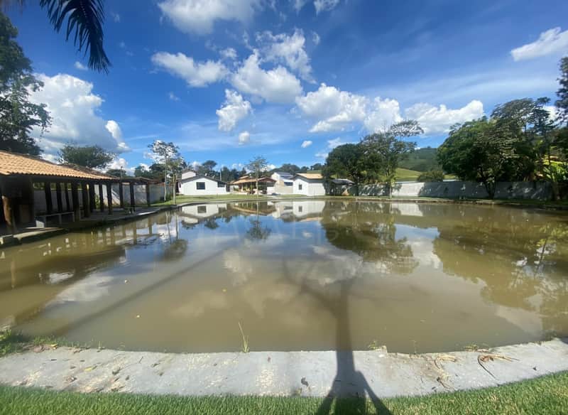 Área externa com lago refletindo casas e céu azul com nuvens. Construções rústicas e gramado.