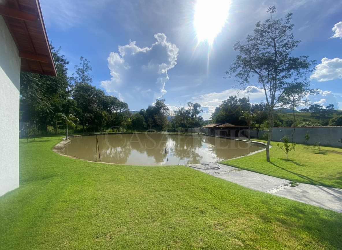 Área externa com lago, gramado verde e construção ao fundo. Céu azul com nuvens e sol forte.