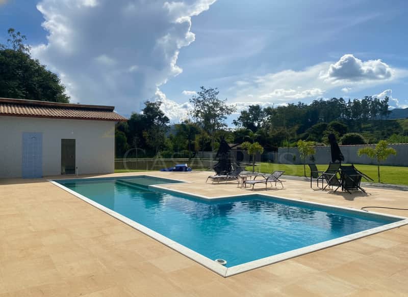 Área externa com piscina azul e espreguiçadeiras em deck bege, sob céu azul com nuvens.