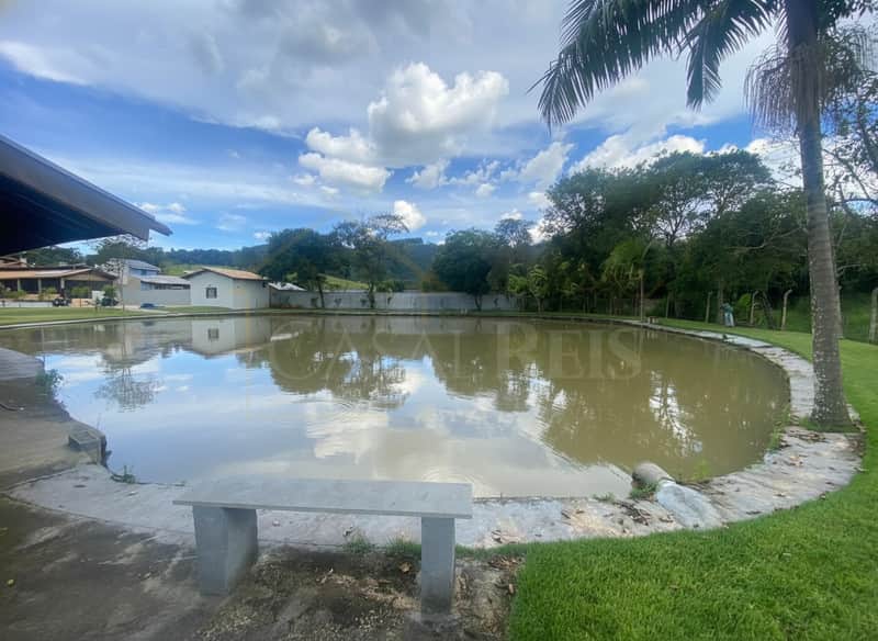 Área externa com lago calmo, gramado verde e banco de concreto. Reflexo de árvores e céu na água.