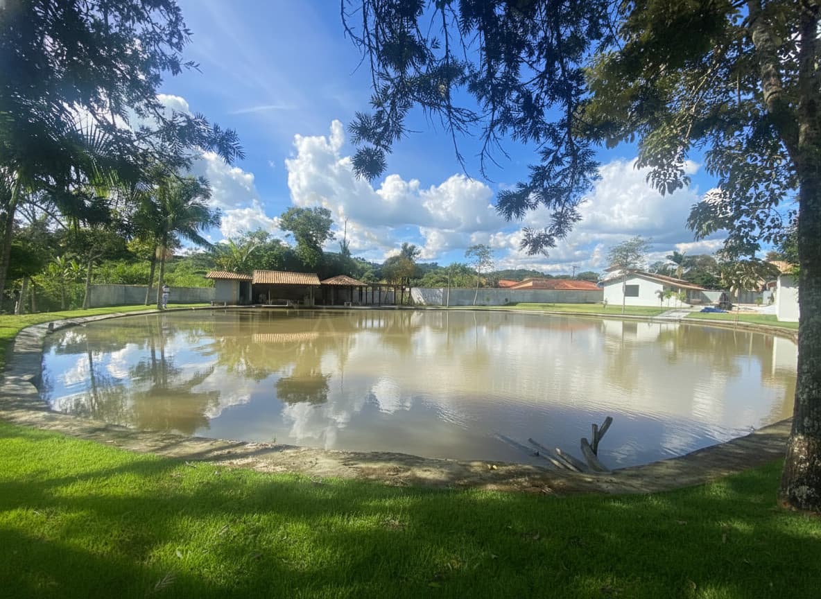 Área externa com lago, gramado verde e quiosques. Reflexo do céu azul na água.