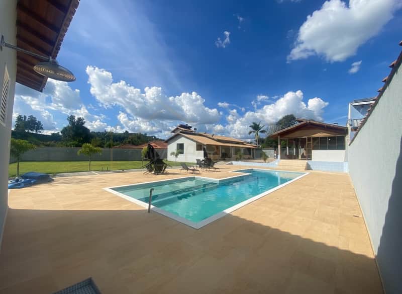 Área externa com piscina azul, espreguiçadeiras e gramado verde sob céu azul com nuvens.