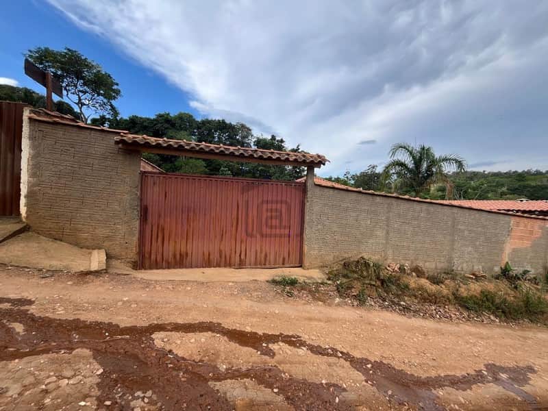 Fachada rústica com portão marrom e muro de tijolos aparentes sob telhado de barro. Terreno irregular.